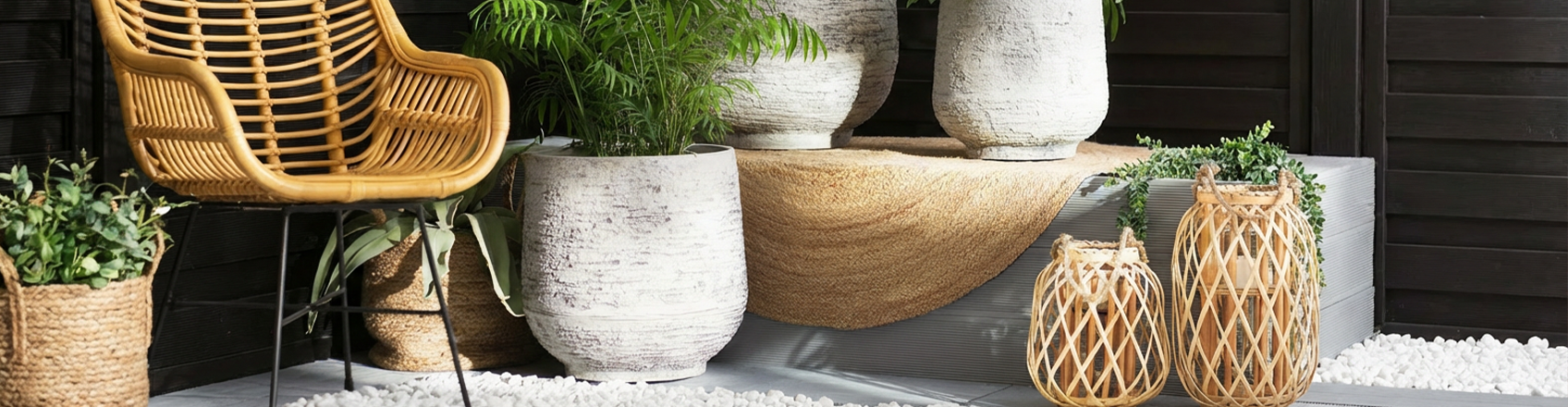 A stylish outdoor patio setup featuring a light rattan armchair, large textured white stone plant pots with lush green plants, and two woven bamboo lanterns on a bed of decorative white pebbles against a dark wooden wall.