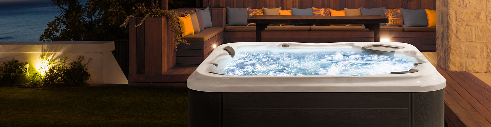 A luxurious outdoor square hot tub illuminated at night with bubbling water, integrated into a wooden deck with a cozy built-in seating area and colorful cushions, overlooking the ocean.