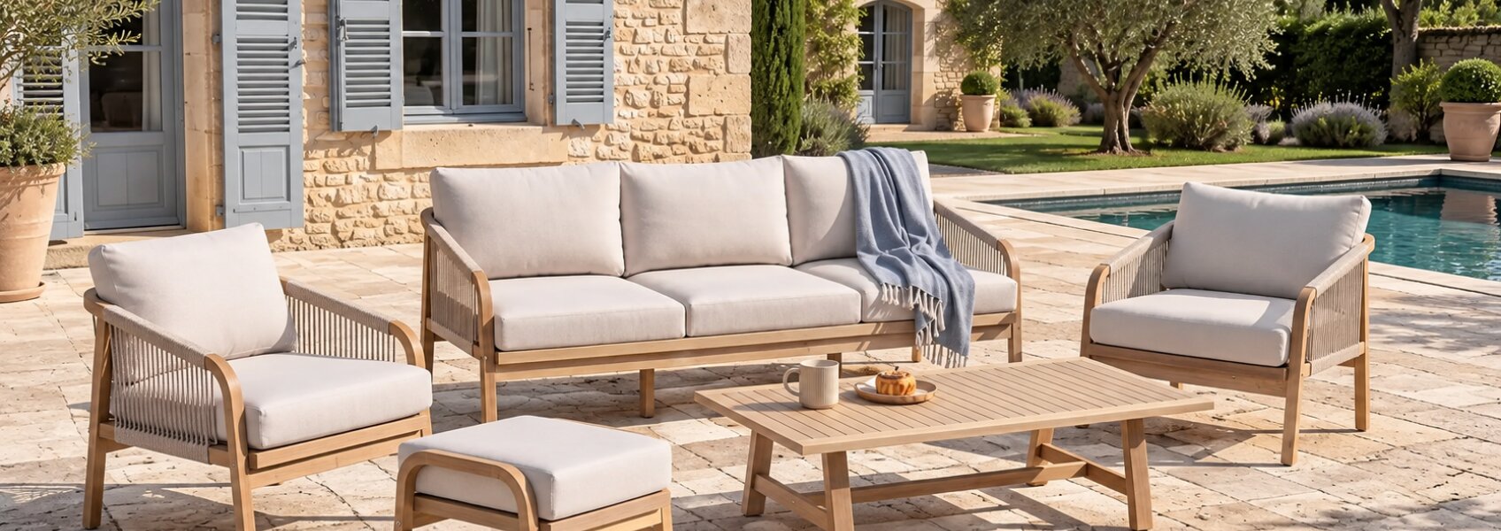 Luxury outdoor lounge set with a teak wood sofa, two armchairs, an ottoman, and a slatted coffee table on a stone patio by a swimming pool.